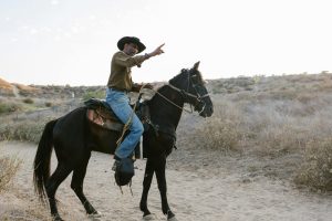 Person on horseback pointing ahead, wearing a cowboy hat and jeans, riding through a dry, brushy landscape.