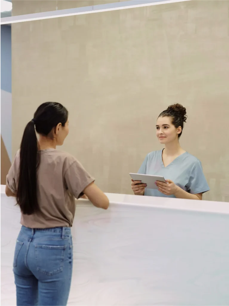 A woman in a blue uniform holds a tablet and stands behind a reception desk, facing another woman with long dark hair wearing a brown T-shirt and jeans.