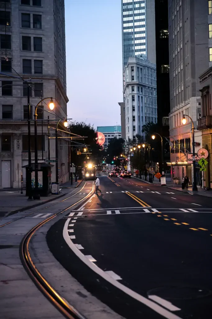 A person crosses an empty city street at dusk, surrounded by tall buildings and illuminated streetlights.