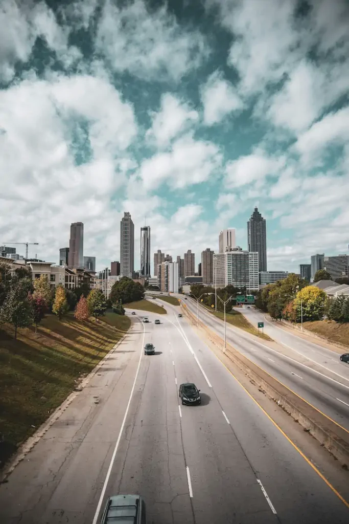 A wide city highway with several cars leads toward a skyline of tall buildings under a partly cloudy sky.