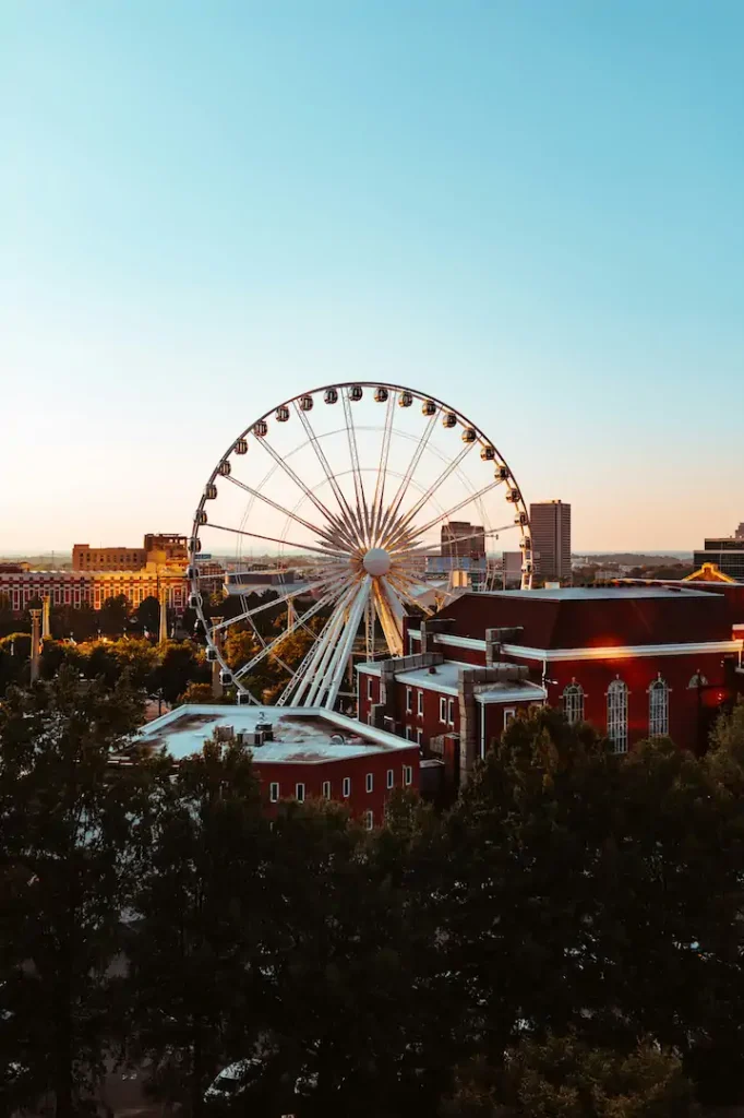 A large Ferris wheel stands behind red brick buildings and trees, set against a clear sky during sunset in an urban area.